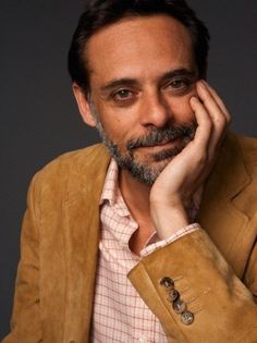 a man sitting down with his hand on his chin and looking at the camera while wearing a brown jacket