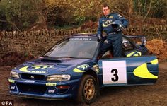 a man standing on the hood of a race car in dirt area next to trees