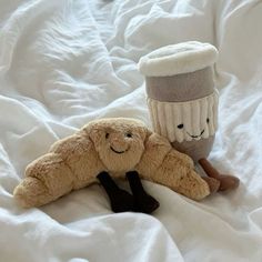 two stuffed animals laying on top of a white bed covered in blankets and pillows with coffee mugs behind them