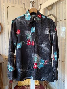 This beautiful shirt is made from a medium weight polyester blend in black, with a white and grey abstract pattern and dark red and teal blue flowers with green leaves. It has a pointy collar, the original buttons on the front and cuffs, and one chest pocket. The measurements, taken with the shirt lying flat, are: shoulder to shoulder, 19 inches; armpit to armpit, 23 inches; sleeves, 25 1/2 inches; length, 30 inches; bottom edge, 23 inches. In very good condition. Retro Black Button-up Blouse, Black Floral Print Shirt For Summer, Black Collared Top With Floral Print, Black Collared Floral Print Top, Black Floral Print Collared Top, Black Printed Collared Blouse, Fitted Black Floral Print Tops, Black Long Sleeve Tops With Abstract Print, Black Vintage Print Top For Summer