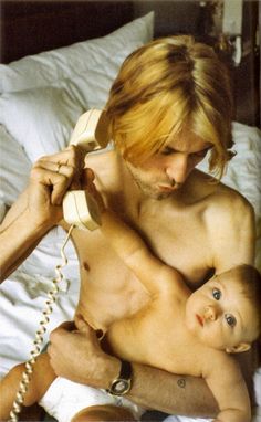 a shirtless man holding a baby while talking on the phone with an adult nearby