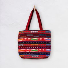 The Akha-Ani large tote bag is the perfect everyday cotton carry-all. It's generous size is designed to hold all the essentials or a lot...! The decoration on the exterior of the bag is hand crafted by the Akha people who live in the hills of  Myanmar. The pattern is colourful and embroidered with different coloured fabrics and threads. *  Bag 46cm Wide x 40cm Depth *  27cm handles *  Inside Bag large open pocket 21cm X 21cm *  Inside zippered pocket 21cm X 21cm *  Also Drawstring compartment in middle of bag *  Cotton with woollen and cotton embroidery *  Padded Base *  Select from a range of woven colour variations Care: Dry Clean only Made in Burma | Fair Trade Disclaimer: Mingalaba products are all made by individual artisans. Each Bag is unique and it's very rare we see 2 perfectly id Red Cotton Canvas Shopping Bag, Red Cotton Canvas Bag For Shopping, Traditional Tote Beach Bag For Travel, Red Cotton Shoulder Bag For Daily Use, Red Square Canvas Bag For Daily Use, Bohemian Red Tote Beach Bag, Red Embroidered Bag For Daily Use, Red Bohemian Beach Bag For Shopping, Embroidered Red Tote Bag