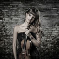 a woman holding a violin in her right hand and posing for the camera with brick wall behind her