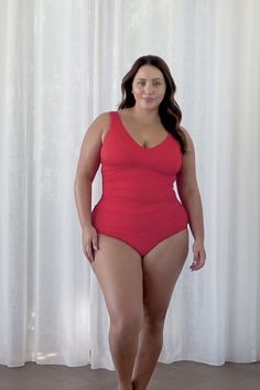 a woman in a red one piece swimsuit standing next to a white curtain with her hands on her hips