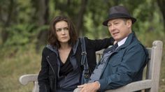 a man and woman sitting on a wooden bench in the woods with trees behind them