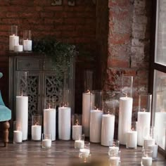 many white candles are lined up on the floor next to a blue chair and brick wall