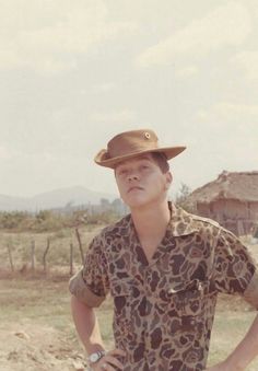 a man wearing a hat standing in front of a dirt field with his hands on his hips