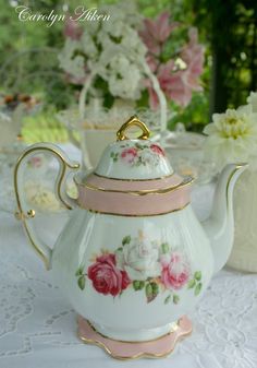 a white tea pot with pink roses on it sitting on a table in front of flowers