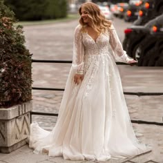 a woman in a long sleeved wedding dress standing on the sidewalk next to a fence