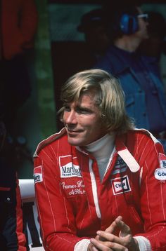 a man with long hair wearing a red jacket and sitting in front of a crowd