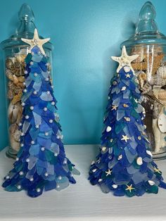 two glass christmas trees sitting on top of a white shelf next to a jar filled with seashells