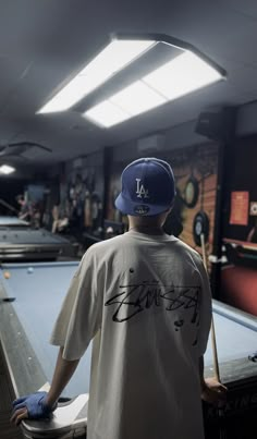 a man standing at a pool table wearing a baseball cap and t - shirt with writing on it