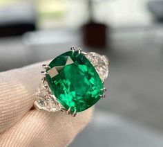 an emerald and diamond ring is being held by someone's hand in front of the camera