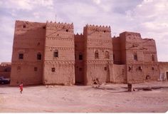 an old building in the middle of a desert with cars parked outside and people walking around