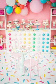 a table and chairs in front of a pink wall with colorful balloons on the ceiling