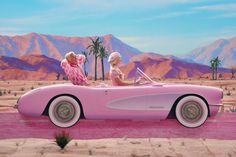 two women sitting in a pink car with palm trees and mountains in the back ground