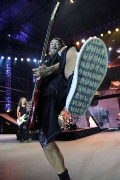 a man holding a guitar while standing on top of a stage with other people in the background