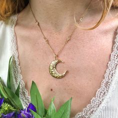 a woman wearing a necklace that has a crescent moon on it and purple flowers in front of her