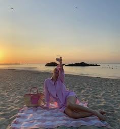 a woman sitting on top of a towel holding a wine glass in her hand while laying on the beach