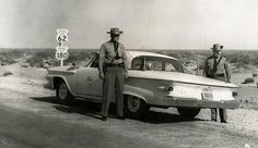 two men standing next to an old car on the side of the road in the desert