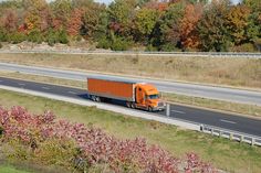 an orange semi truck driving down the highway in autumn time with trees and grass around it