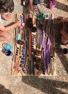 several children sitting on the floor and one is painting some pine cones with purple paint