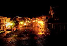 an empty city street at night with lights on and cars parked in the dark streets