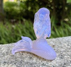 a glass mermaid figurine sitting on top of a stone slab in front of some trees