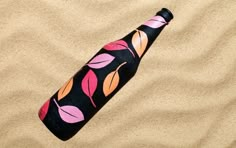 a bottle on the sand with pink and orange leaves painted on it's side