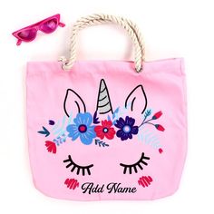 a pink tote bag with an unicorn face and flowers on it, next to sunglasses