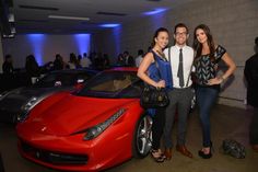 two women and a man standing next to a red sports car in a room full of people