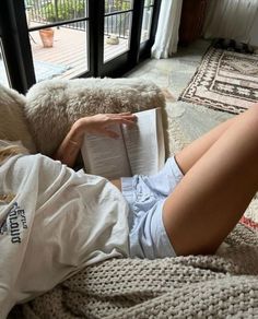 a woman laying on top of a bed next to a window with a book in her hand