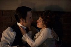 a man and woman sitting next to each other in front of a brick wall with their arms around each other