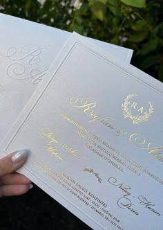 a person holding up two white and gold wedding cards in front of some trees with green leaves