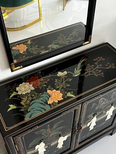 an ornate black and gold painted dresser with flowers on it next to a large mirror