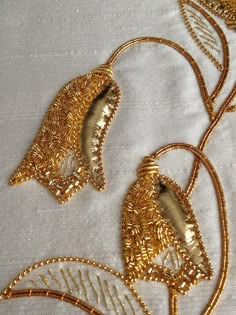 two gold earrings are sitting on a white tablecloth with golden thread and beading