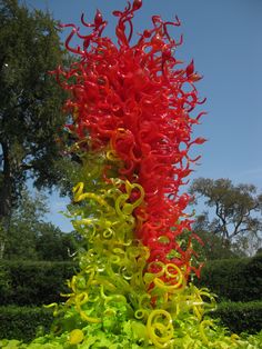 a tall red and yellow glass sculpture in the middle of a garden with trees behind it