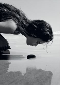 a woman kneeling down on top of a sandy beach next to the ocean with her head in the sand