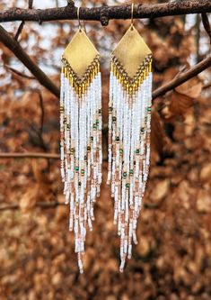 two gold and white earrings hanging from a tree branch in front of some brown leaves