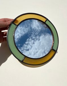 a hand holding up a round mirror with clouds in the sky reflected in it's side