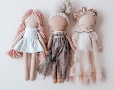 three dolls are sitting next to each other on a white surface, one is wearing a dress and the other has pink hair