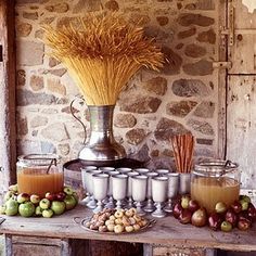 an assortment of fruits and drinks on a table in front of a stone wall with a vase filled with wheat