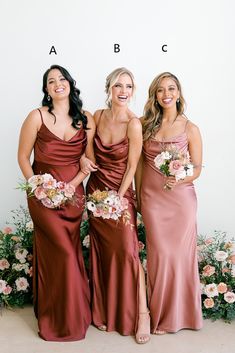 three women in long dresses standing next to each other and smiling at the camera with flowers behind them