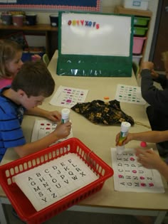 three children sitting at a table working on letters and numbers with cups in front of them