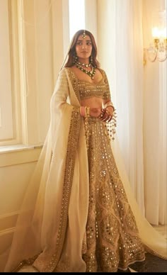 a woman standing in front of a window wearing a gold and white lehenga