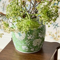 a green vase filled with lots of flowers on top of a wooden table next to a wall