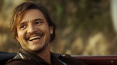 a man with long hair smiling and wearing a brown shirt in front of a red car