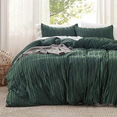 a bed with green bedspread and pillows in front of a white curtained window