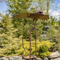 a metal bird sculpture sitting on top of a wooden stand in front of some bushes