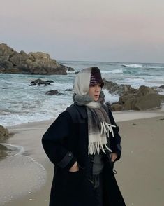 a woman standing on top of a sandy beach next to the ocean wearing a hat and scarf